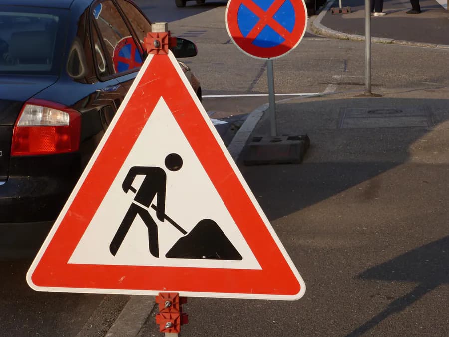 Verkehrszeichen 123 „Arbeitsstelle“ auf mobilem Ständer am Straßenrand, dahinter ein Haltverbotsschild auf Fußplatte