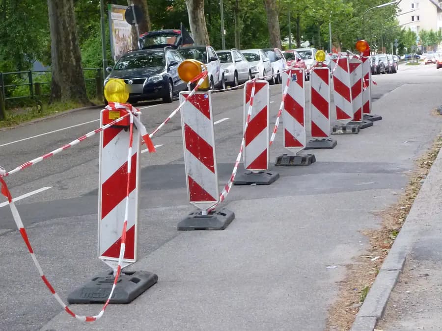 Gelbe Warnleuchten auf rot-weißen Leitbaken mit Fußplatte, verbunden mit Absperrband am Fahrbahnrand